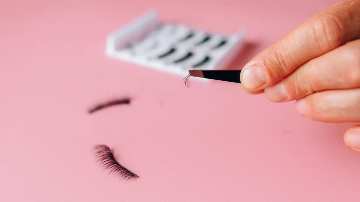Close-up of applying false eyelashes with tweezers over a pink background.