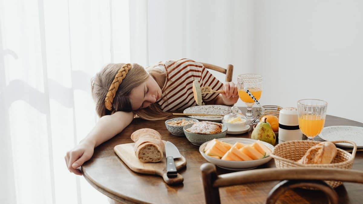 Young girl dozing at breakfast table with diverse food spread, amusing morning scene.