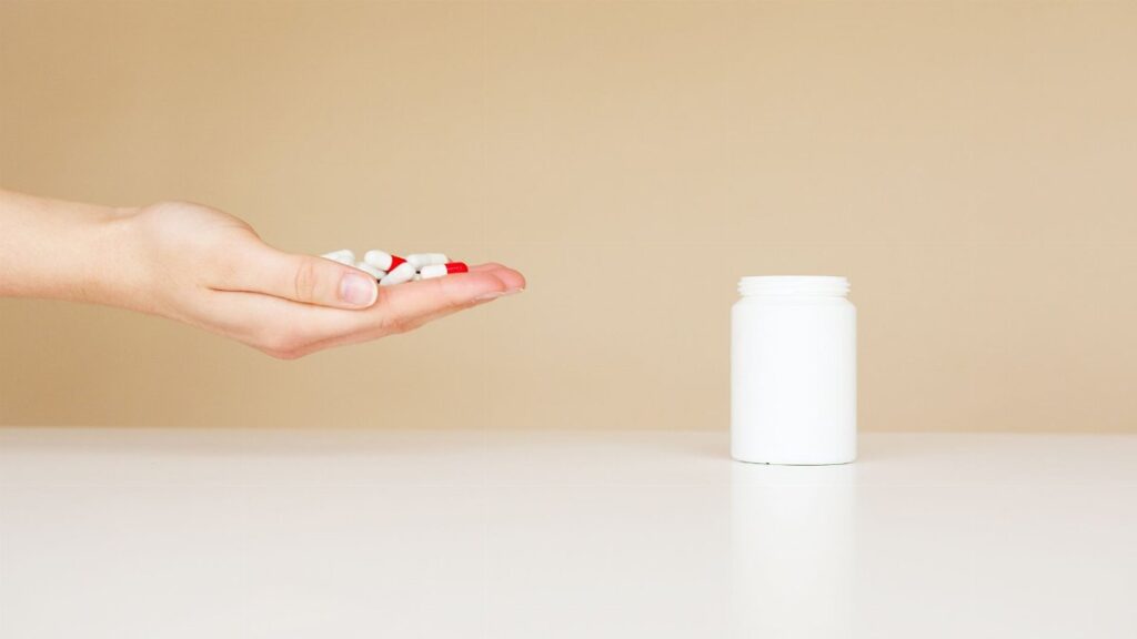 Side view of crop unrecognizable person with palm full of double colored pills near table with white plastic jar