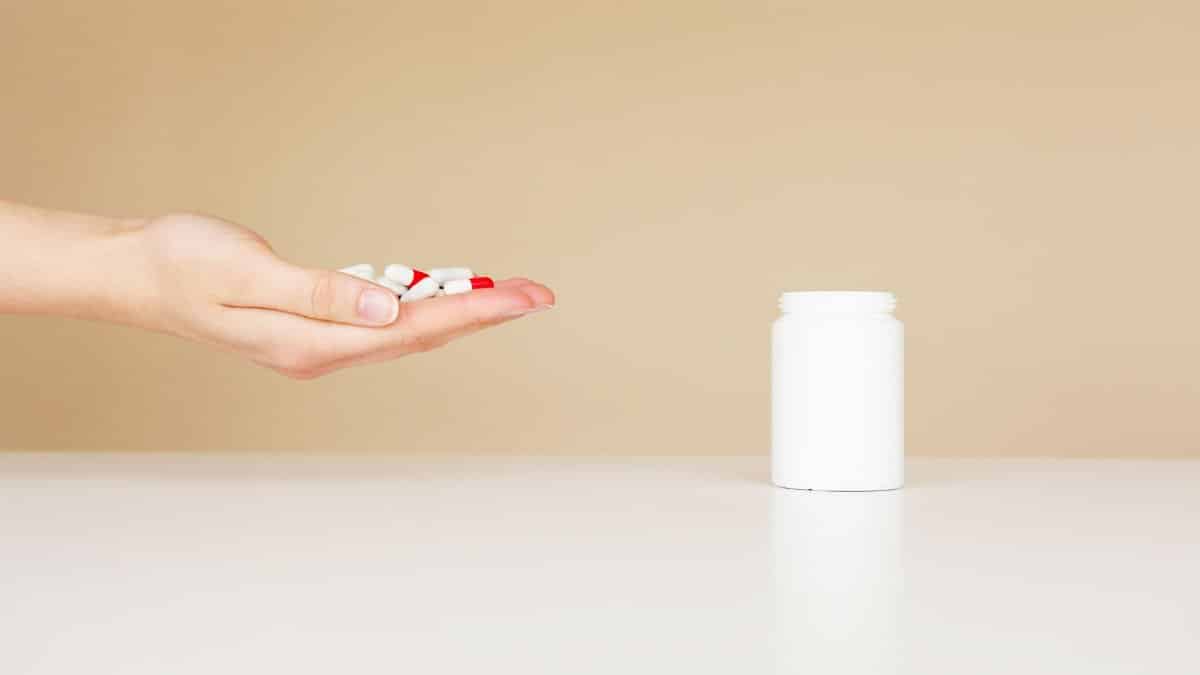 Side view of crop unrecognizable person with palm full of double colored pills near table with white plastic jar