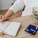 Adult holding cash and writing in planner while using a calculator at home.