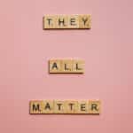 Wooden Scrabble tiles spell 'They All Matter' on a pink background, promoting inclusivity.