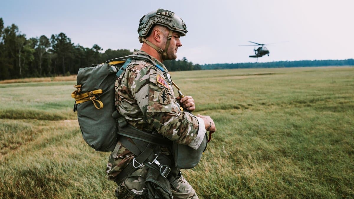 A soldier in camouflage walking in a field with a helicopter overhead.