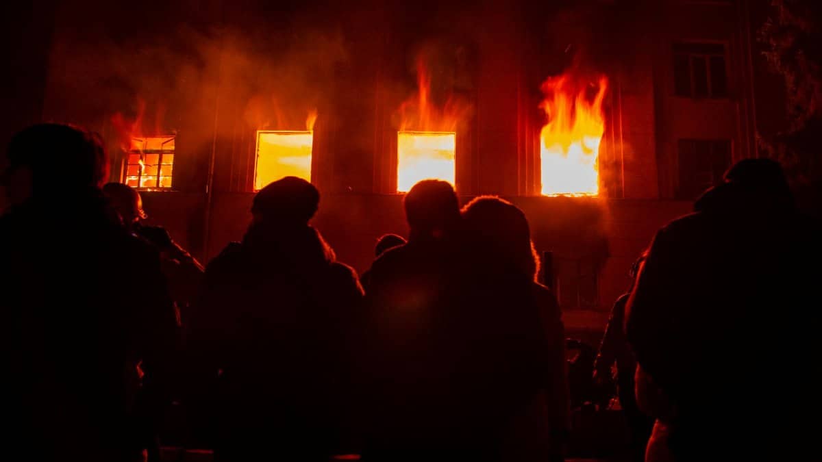 Silhouetted figures watch a building engulfed in flames at night, dramatic firelight contrasts with the dark.