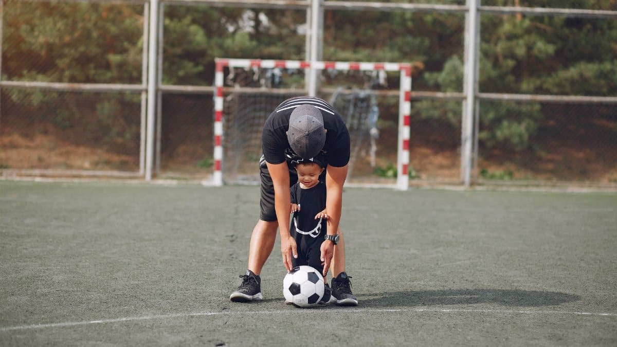 Full body anonymous father in black casual wear and toddler son walking with football ball on green sports ground on sunny day