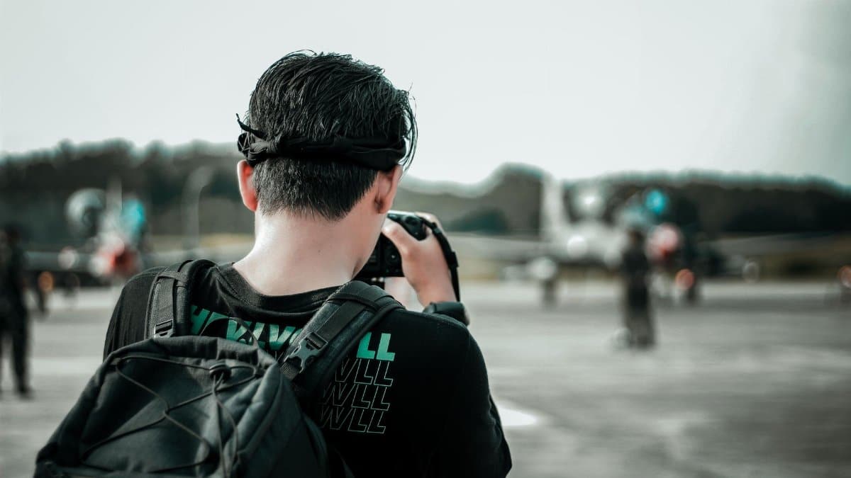 A man with a backpack photographs airplanes on an outdoor runway during the day.