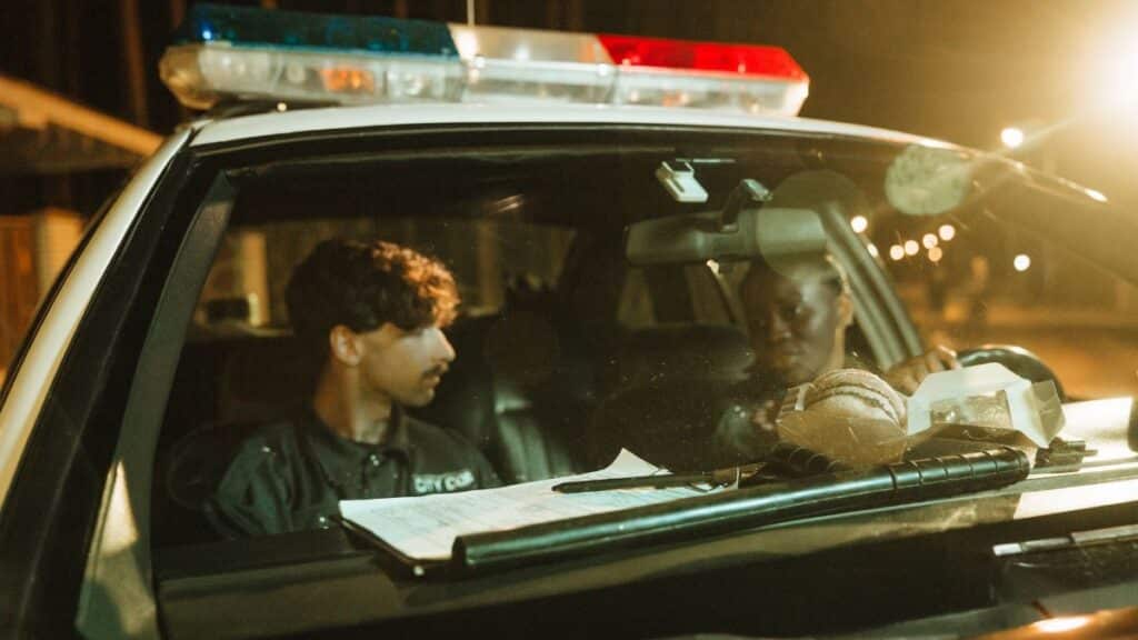 Two police officers in a patrol car during the night, engaging in a conversation.