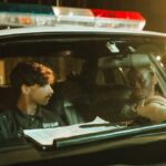 Two police officers in a patrol car during the night, engaging in a conversation.