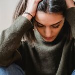 Crop disappointed young ethnic female with dark hair in casual clothes grabbing head with hands and looking down while sitting against white wall at home