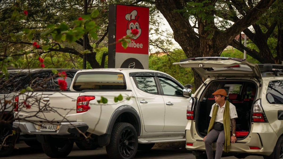 A man in an open car trunk at Manila parking lot with Jollibee sign nearby.
