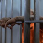 A tattooed inmate's arms rest on jail cell bars, wearing an orange uniform inside a prison.