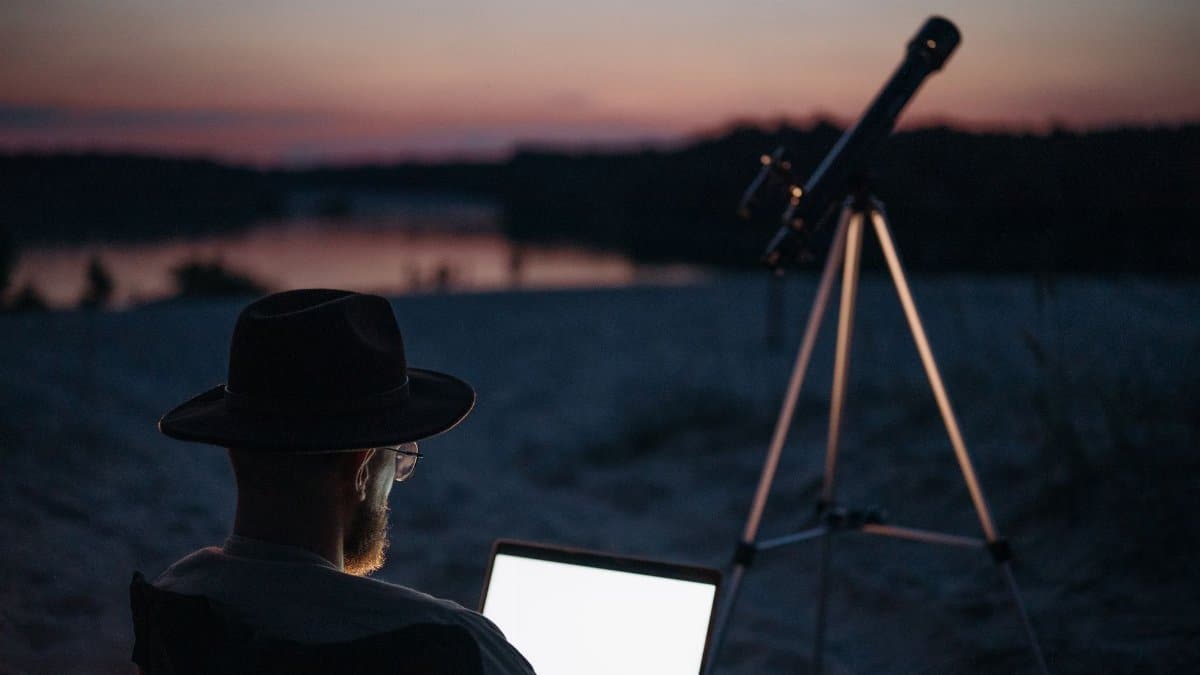 A man enjoying stargazing by a serene lake at twilight, using a telescope and laptop.