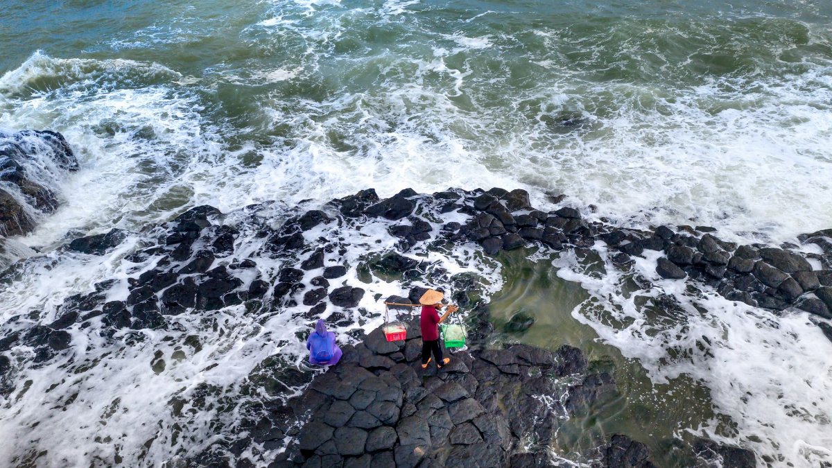 Two individuals on a rocky coast amid crashing waves observed from above.