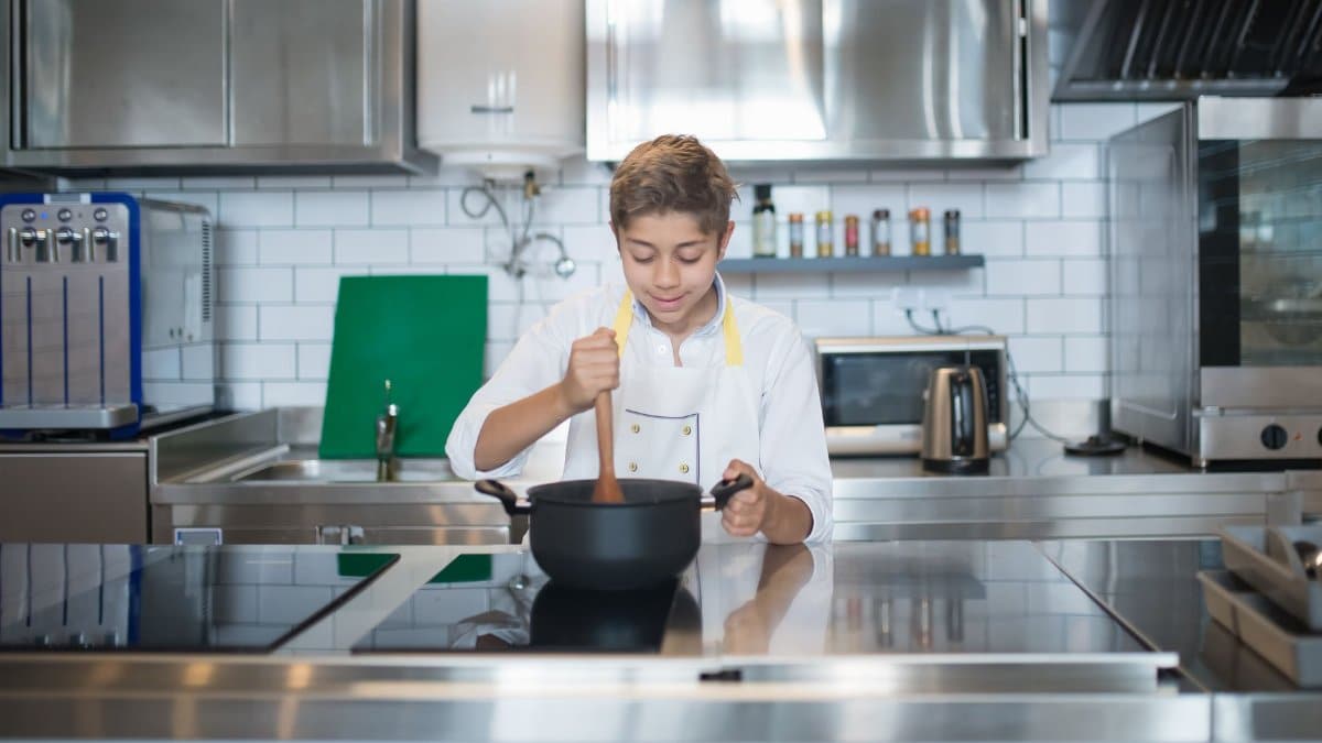 An aspiring young chef in an apron cooks with focus in a sleek, modern kitchen setting.