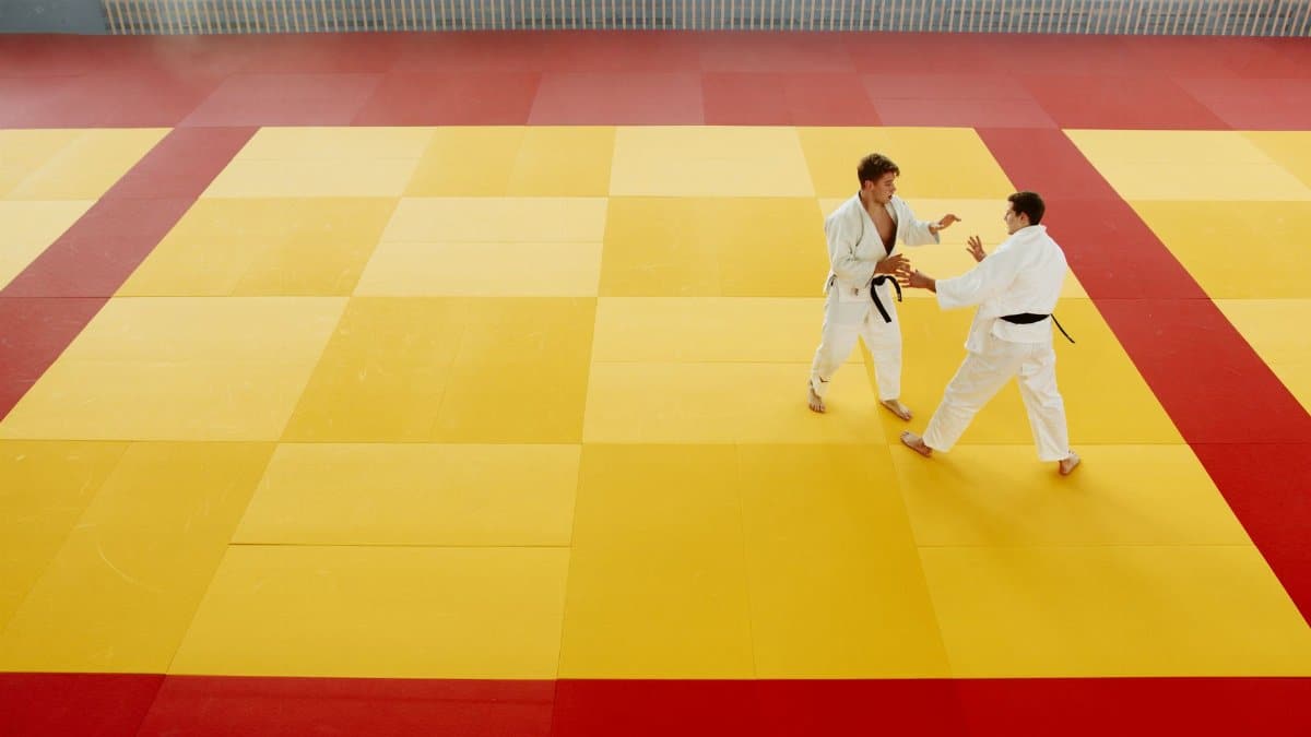 Two martial artists practice techniques on a yellow and red mat in an indoor gym.
