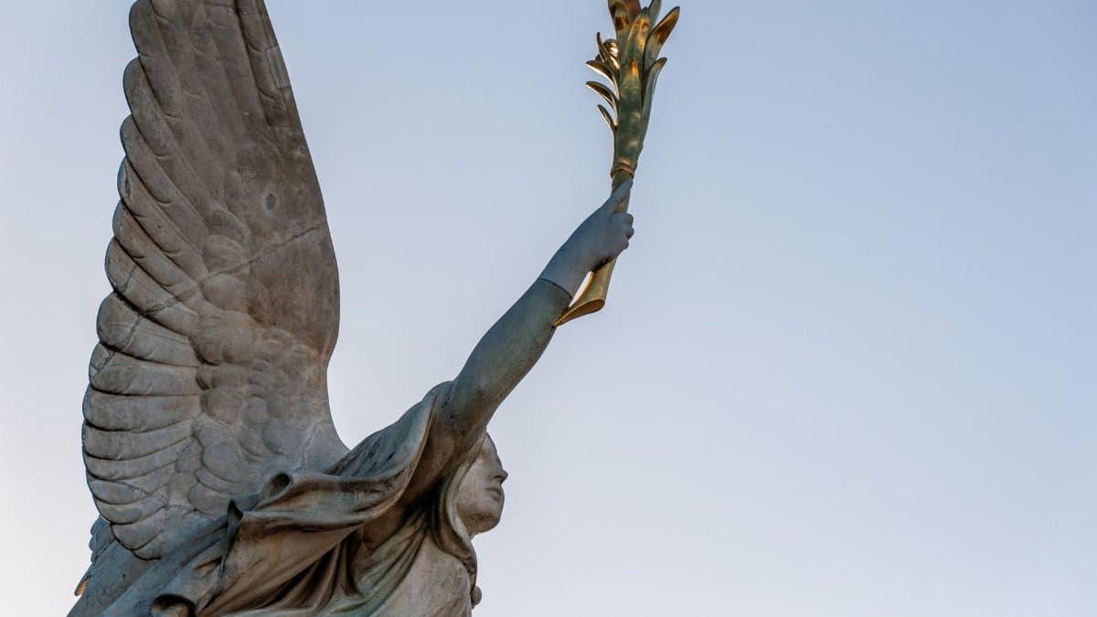 A majestic angel statue holding a golden palm leaf reaches towards the clear blue sky.