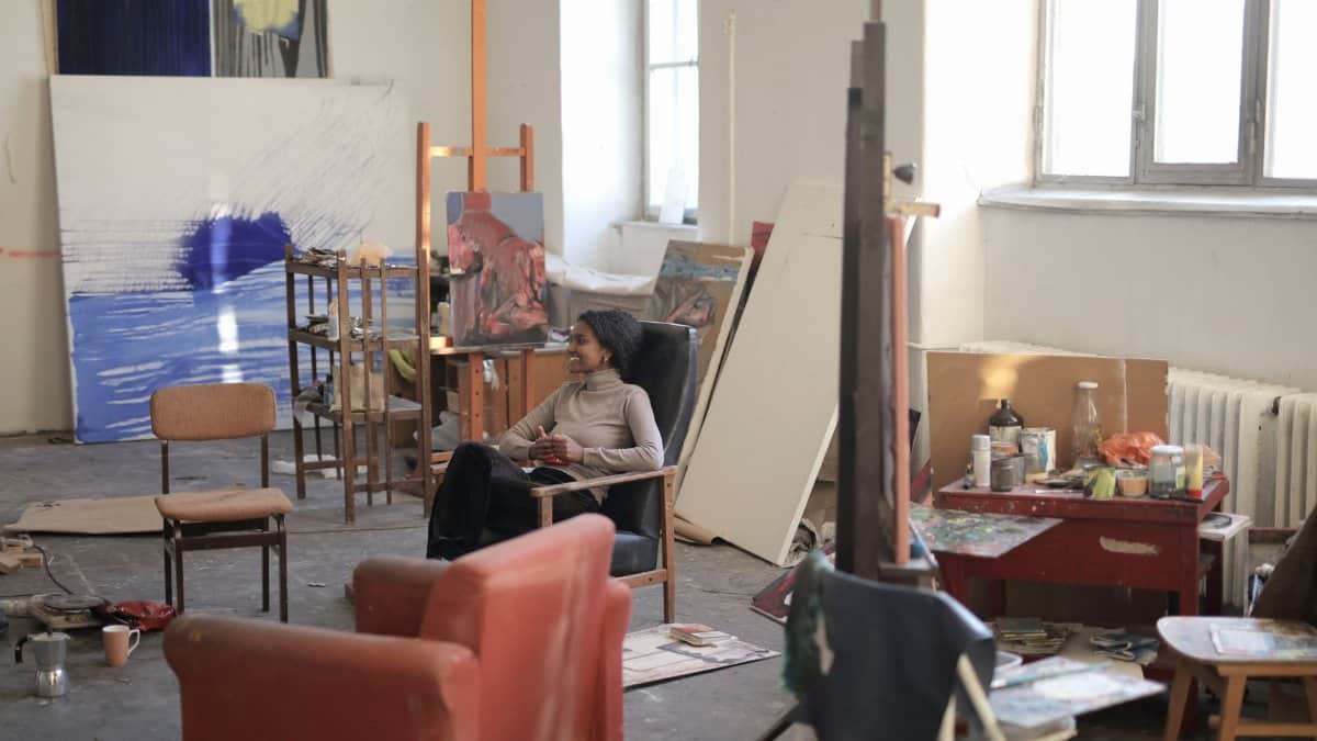 A creative and cozy art studio with a relaxed artist sitting amidst paintings and art supplies.