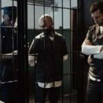 A police officer stands by two prisoners in jail uniforms behind bars, indoors.