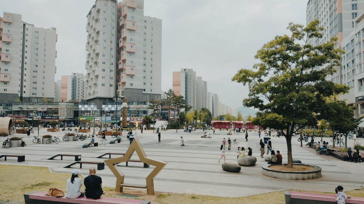 Bustling urban park in Hanam-si, South Korea with people relaxing among modern high-rise buildings.