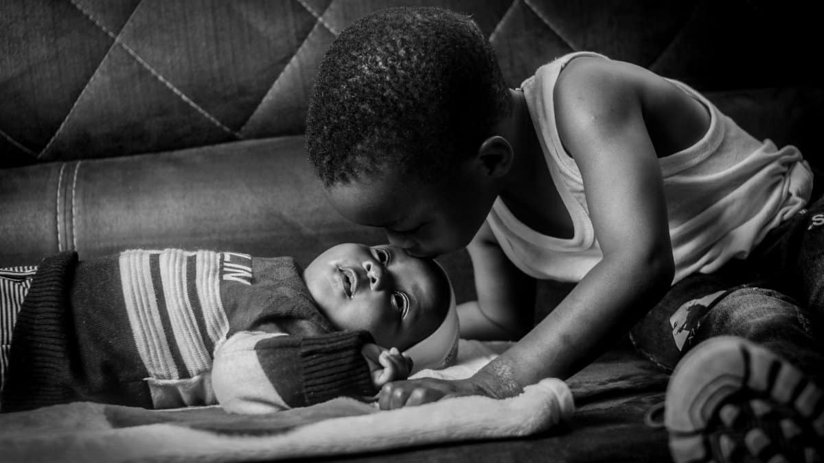 Touching black and white photo of a young boy kissing his baby sibling indoors.