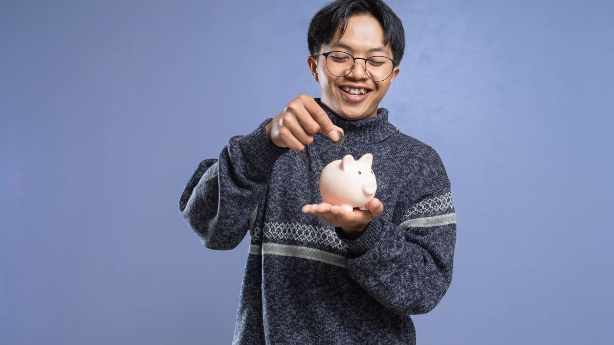 Smiling man holding a piggy bank and inserting a coin, representing savings and financial planning.