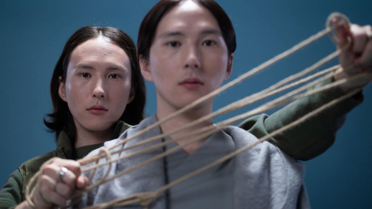 Two young men engaged in a conceptual pose with ropes against a blue backdrop, exploring connection and tension.