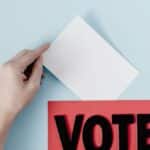 A hand places a ballot into a red box with the word VOTE against a blue background.
