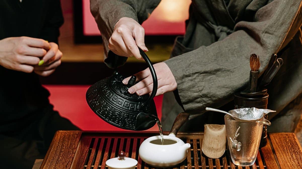 Close-up of a traditional tea ceremony showcasing hands pouring tea into a ceramic teapot on a wooden table.