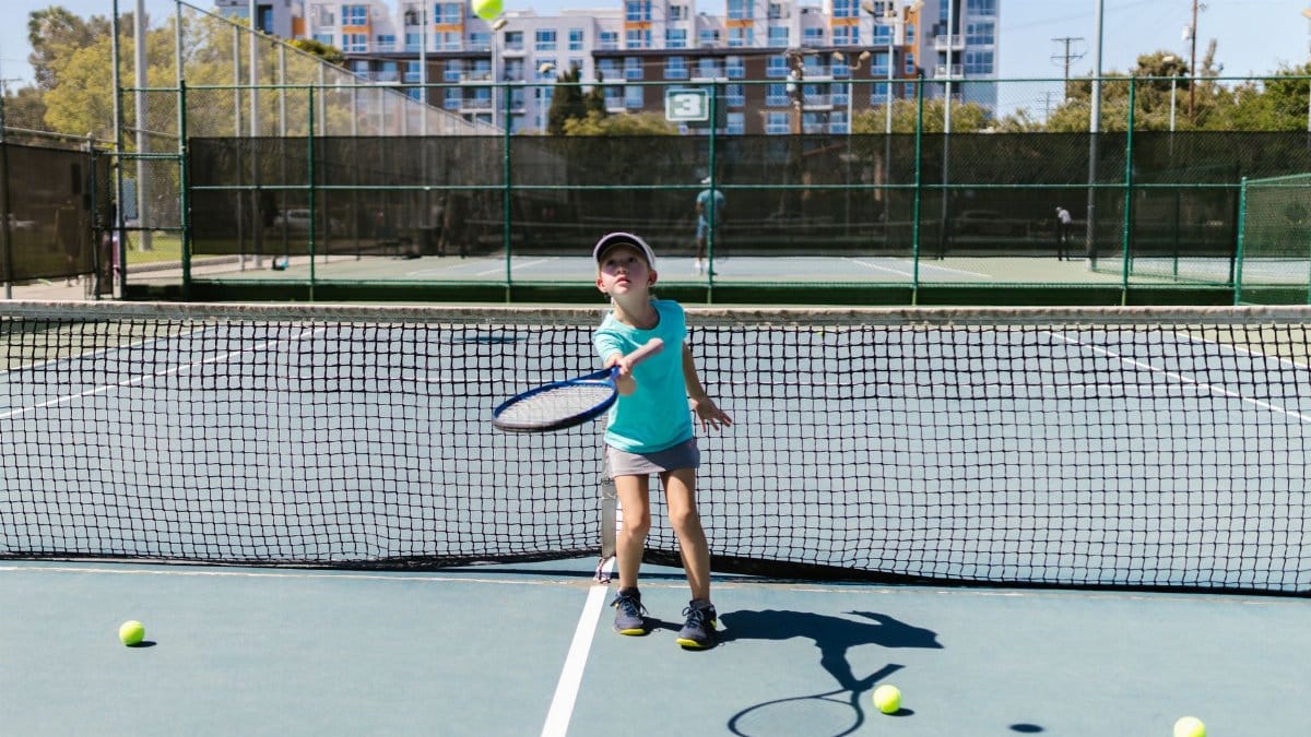 A young girl in sportswear practices tennis on a sunny outdoor court with multiple tennis balls around.