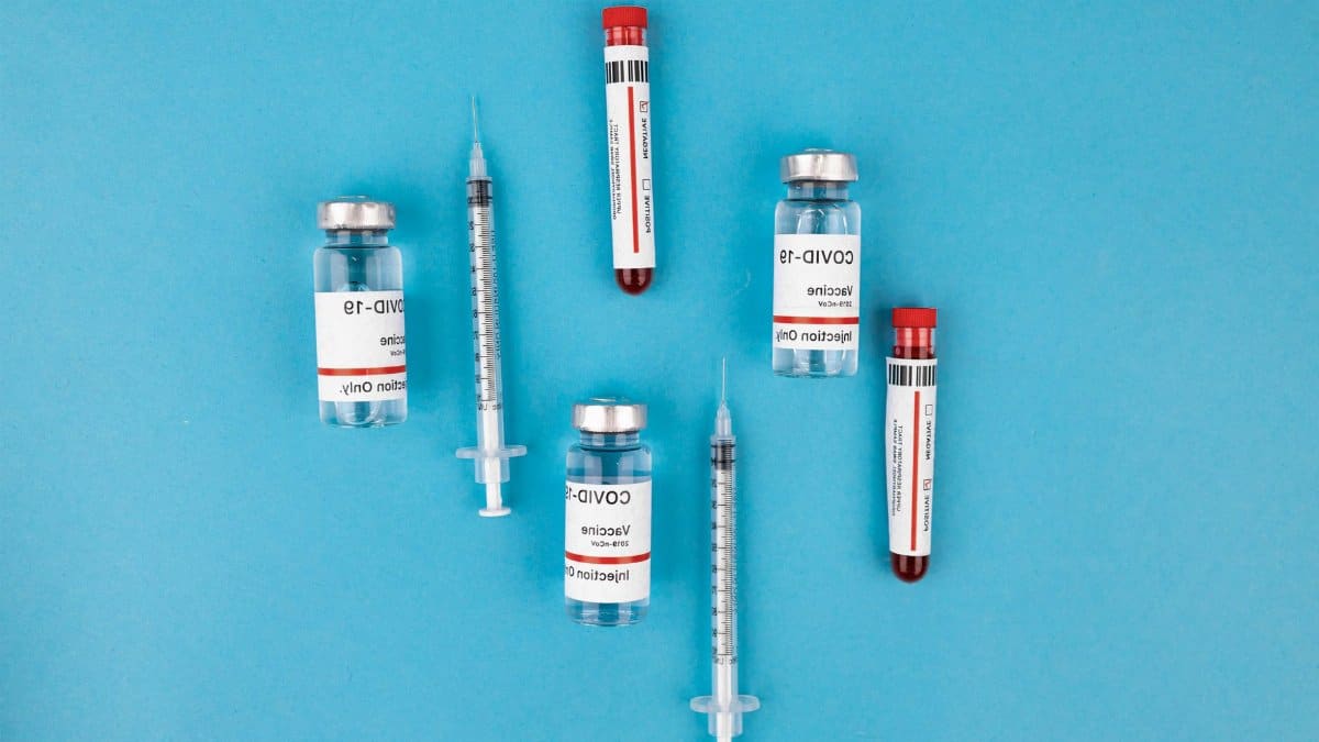 Top view of COVID-19 vaccine vials, syringes, and test tubes on a blue background, representing immunization and protection.