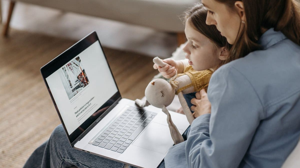 A mother works from home on a laptop with her daughter sitting on her lap, holding a stuffed toy.