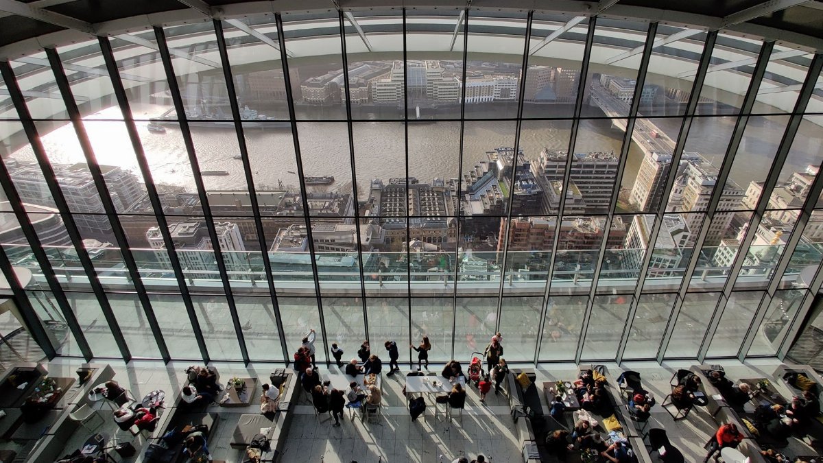 People inside a modern atrium with views of London's skyline and river during daytime.