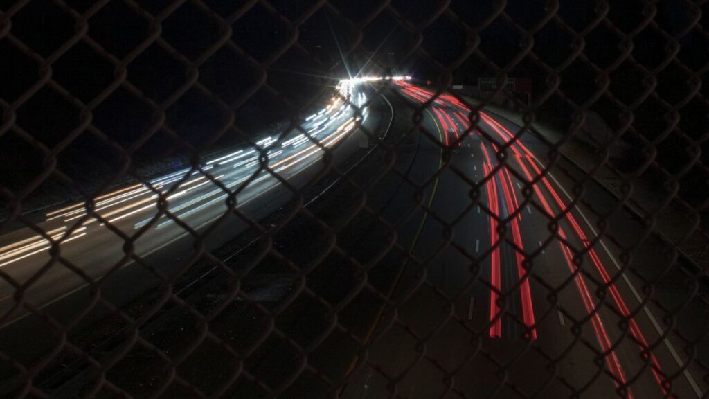Long exposure capture of car light trails through a chain link fence at night.