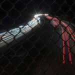 Long exposure capture of car light trails through a chain link fence at night.