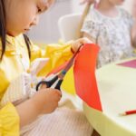 A young girl cuts colorful paper during an arts and crafts activity in a preschool classroom.