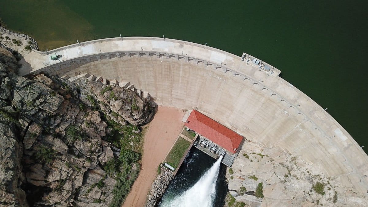 Aerial shot of a hydroelectric dam releasing water into the river below, surrounded by rocky terrain.