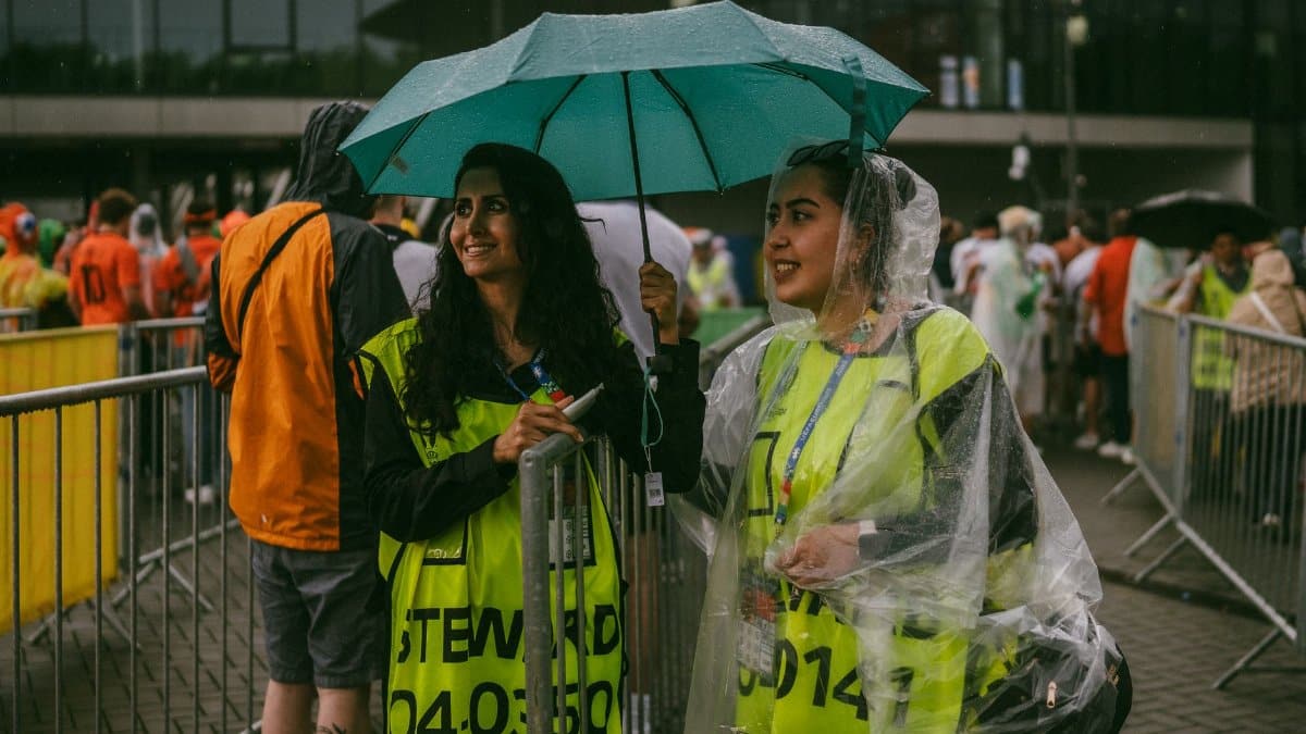 Two female stewards smiling under an umbrella at an outdoor event in rainy weather