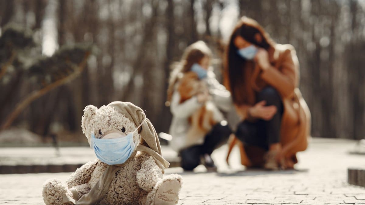 Cute children toy in scarf and mask placed on asphalt against blurred owners woman and girl in warm wear playing in nature
