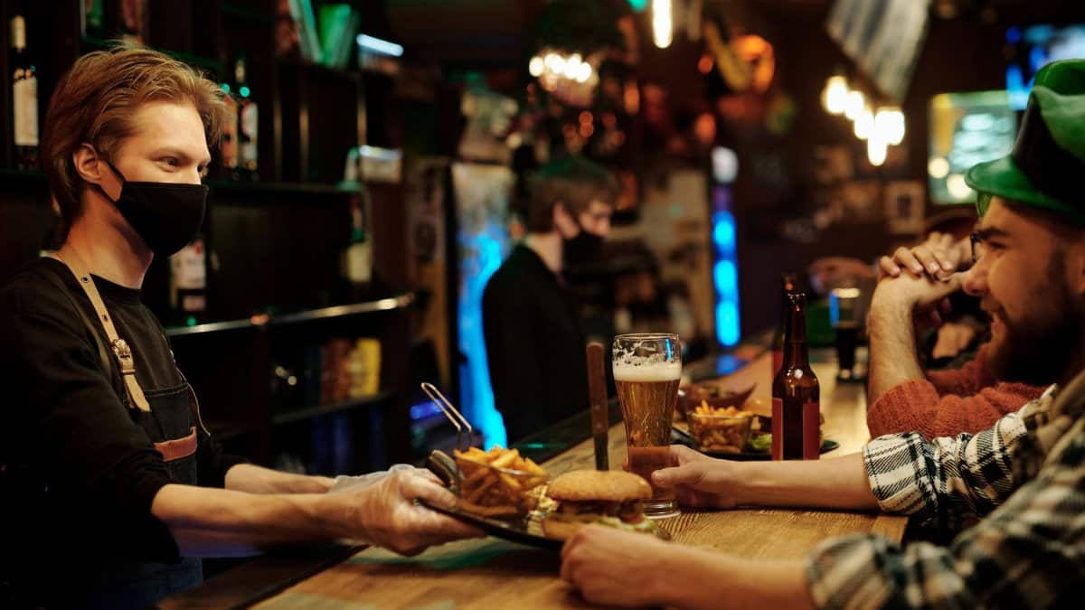 Bartender with mask serving food and drinks to patrons in a lively pub setting.