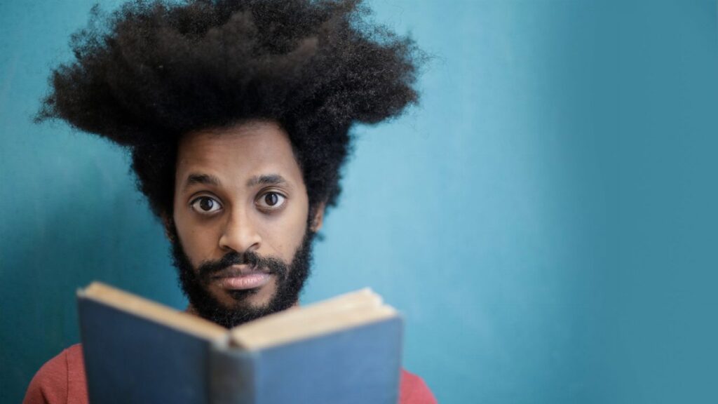 Portrait of a man with afro hair reading a book against a blue background, expressing focus and wisdom.