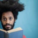 Portrait of a man with afro hair reading a book against a blue background, expressing focus and wisdom.