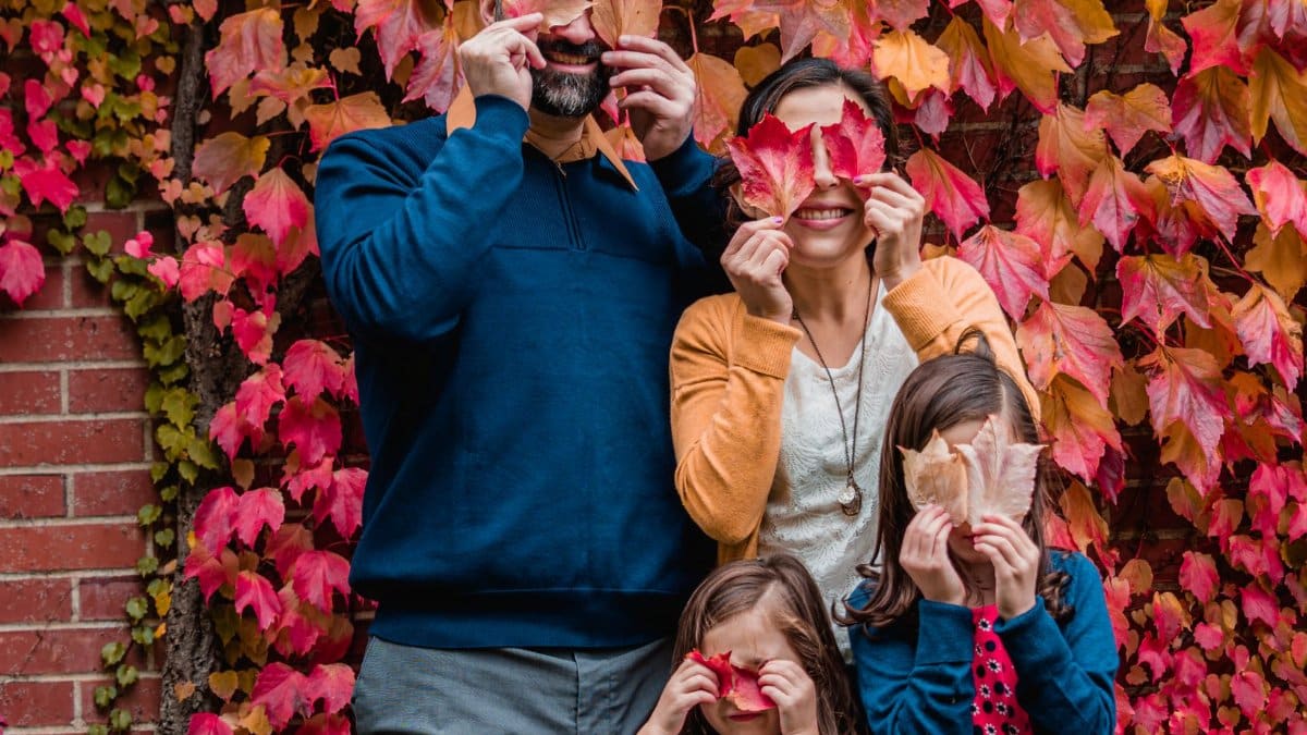 A family of four playfully holding autumn leaves in front of their faces against a colorful leaf backdrop.