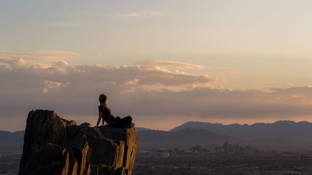 A woman sits on a rock formation overlooking Phoenix, Arizona at sunset, embracing nature and tranquility.