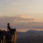 A woman sits on a rock formation overlooking Phoenix, Arizona at sunset, embracing nature and tranquility.