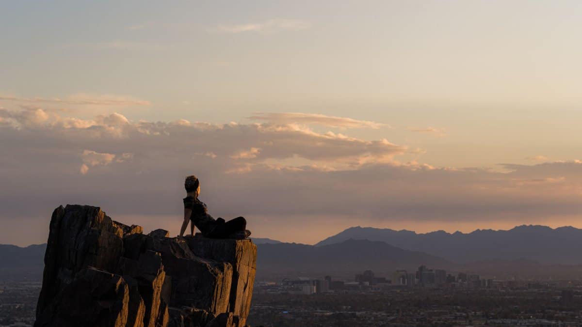 A woman sits on a rock formation overlooking Phoenix, Arizona at sunset, embracing nature and tranquility.