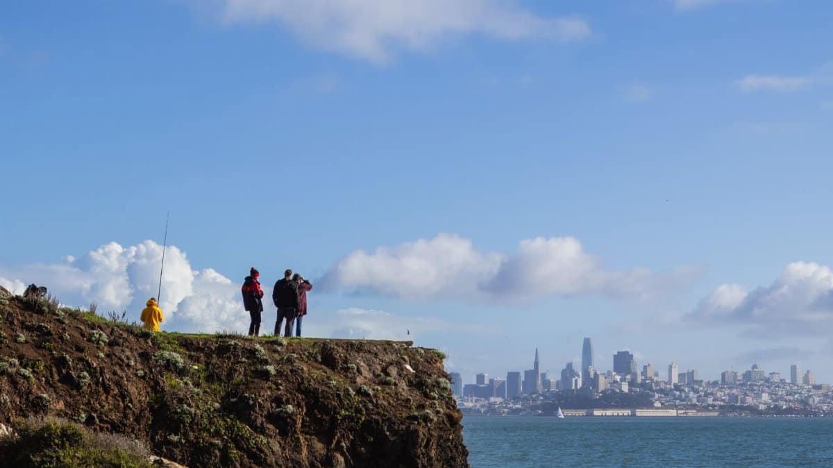 Tourists enjoying a view of San Francisco skyline from a rocky coast under a clear blue sky.