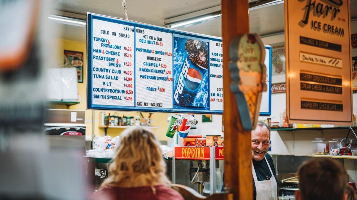 A retro diner setting with a vintage menu board, customers, and a smiling staff member.