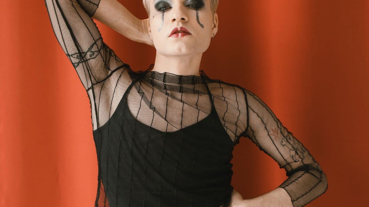 Striking portrait of a stylish individual with bold makeup and sheer attire, against a vibrant red backdrop.