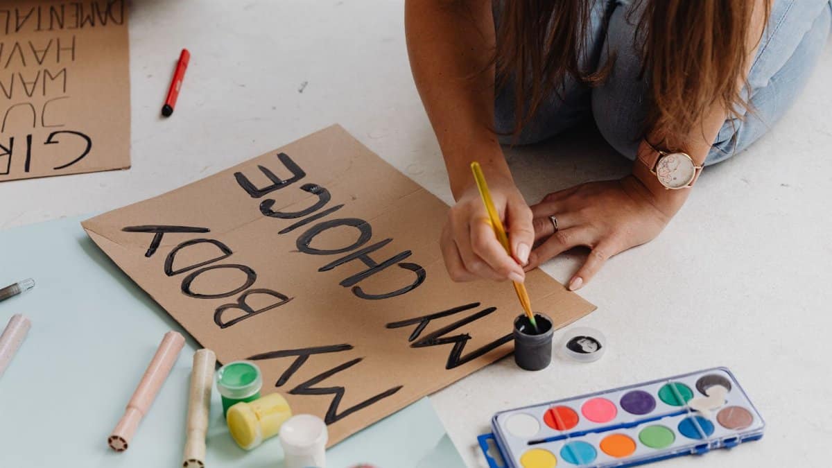 Woman preparing protest sign with empowering message indoors, promoting women's rights.