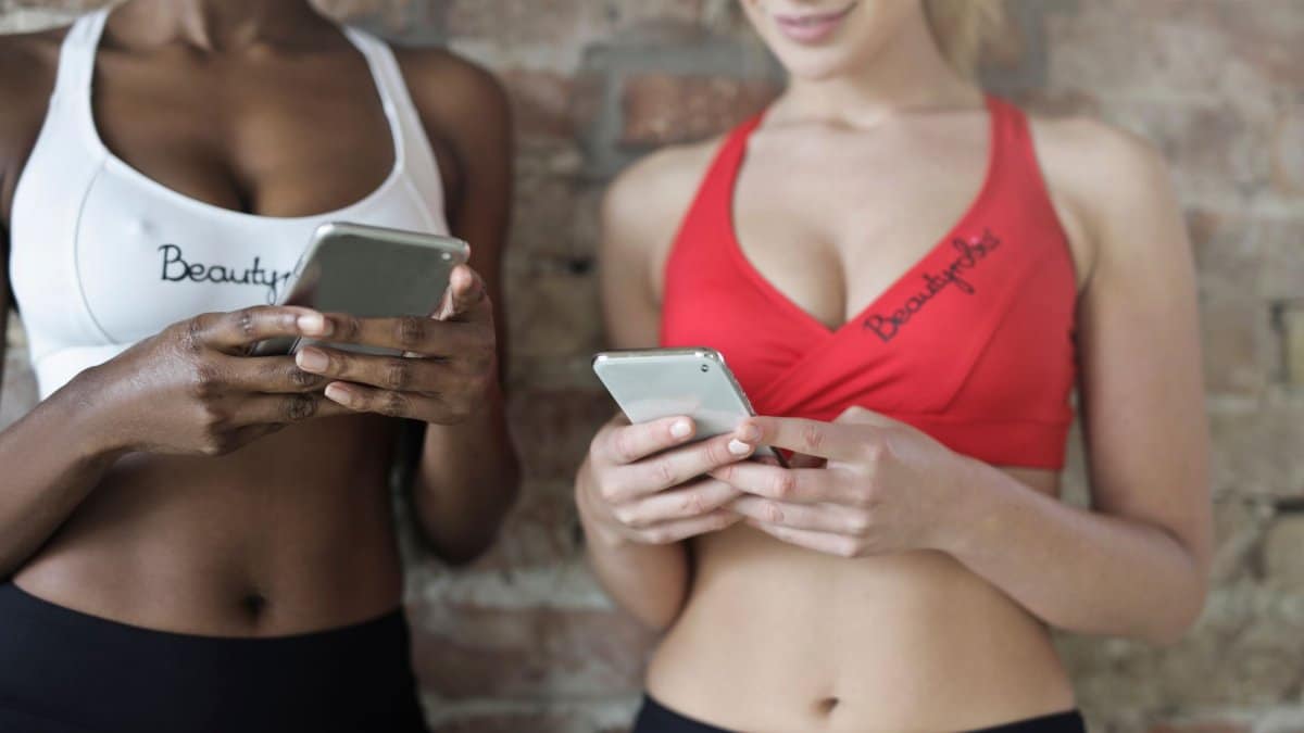 Close-up of two women in athletic wear using smartphones against a brick wall.